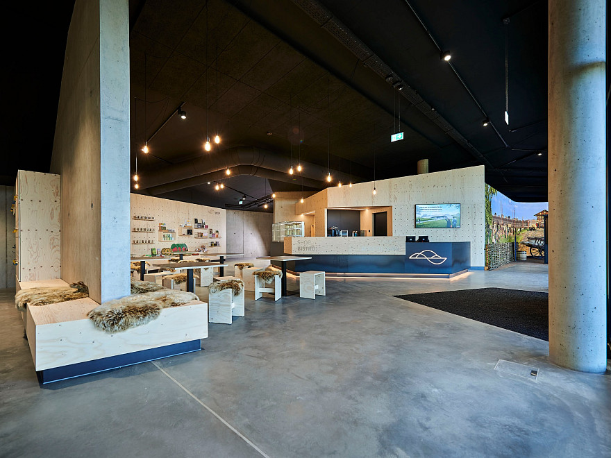 Heller Eingangs- und Empfangsbereich eines modernen Besucherzentrums mit Sitzgelegenheiten aus hellem Holz, Verkaufsregalen, Empfangstheke und großzügigem Betonboden. Im Hintergrund führen Zugänge zu den Ausstellungsbereichen.