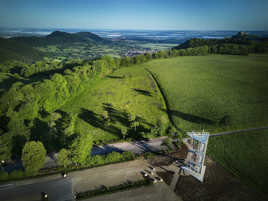 Ein Luftbilds eines modernen gestalteten Aussichtsturms aus Metall und Holz vor der beeindruckenden Kulisse der vorderen Alb einer grünen Wiese, die direkt auf den Albtraum zuläuft und dann abrupt abbricht im Hintergrund den Ausblick auf tiefer liegende Ortschaften und auslaufende Berge.
