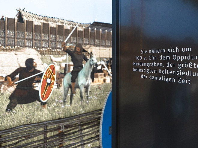 Ein Bildschirm zeigt eine digitale Rekonstruktion des Oppidums Heidengraben mit keltischen Kriegern vor einer befestigten Siedlung. Daneben informiert ein Text über die größte befestigte Keltensiedlung ihrer Zeit.