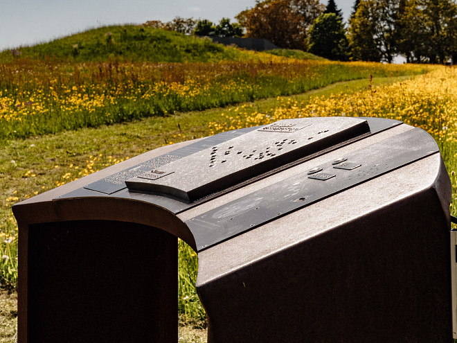 Eine bronzene Installation mit Informationen auf einer gelbblühenden Wiese, im Hintergrund ein keltischer Grabhügel.