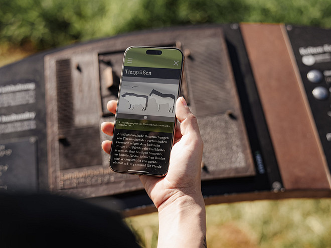 Eine Hand, die ein Handy hält und auf dem Bildschirm sind Informationen zu alten Nutztieren abgebildet, im Hintergrund eine bronzene Skulptur als Platte.