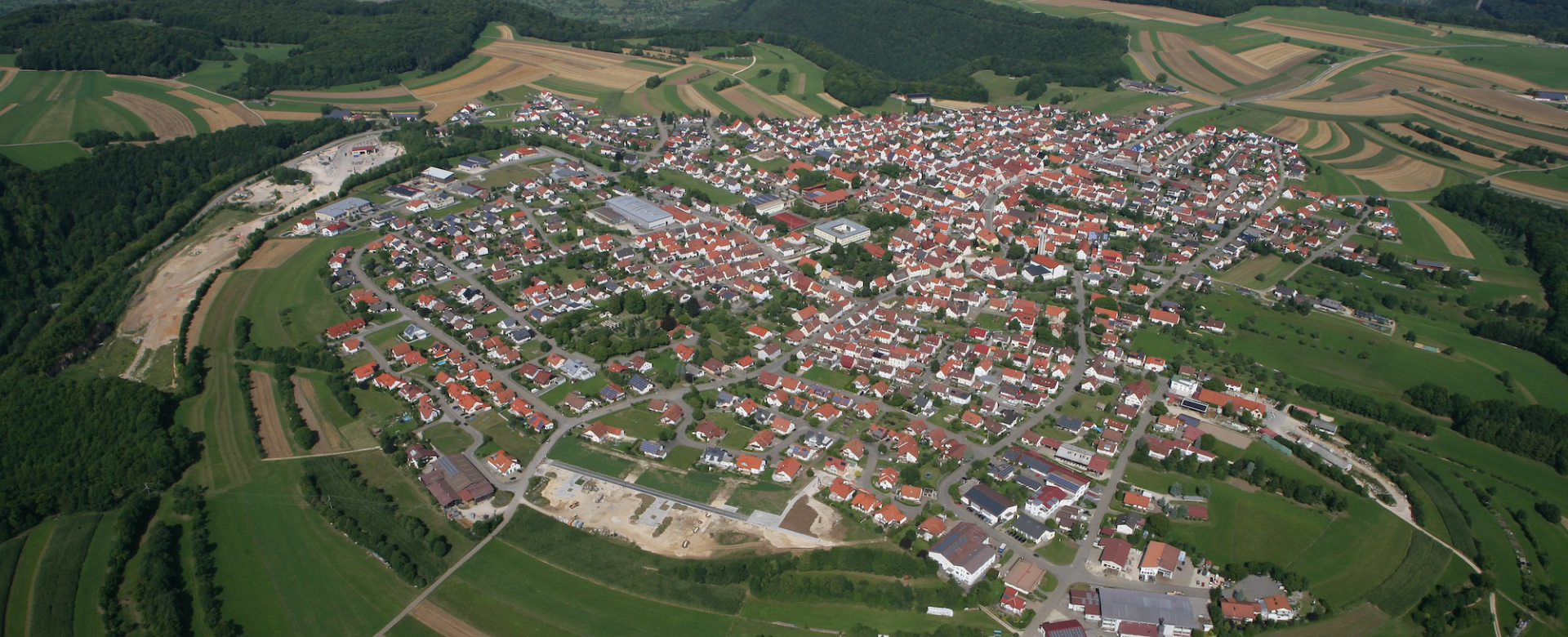 Eine Luftaufnahme zeigt den Ort Hülben mit roten Ziegeldächern, umgeben von landwirtschaftlichen Flächen, Wäldern und den Hügeln der Schwäbischen Alb.