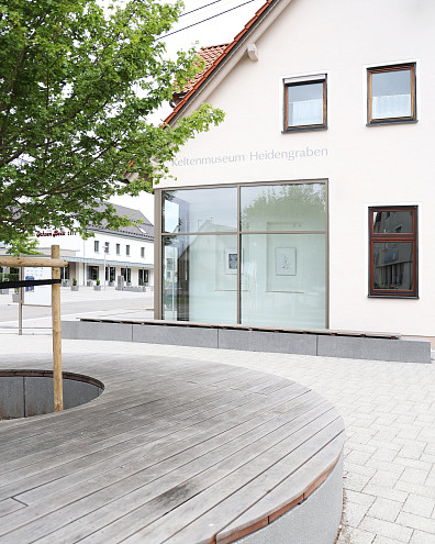 Im Vordergrund eine runde Bank um einen Baum herum, halb abgeschnitten im Bild, im Hintergrund ein Haus mit modernem glasartigen Eingangsbereich. Auf dem Haus steht "Keltenmuseum Grabenstetten".