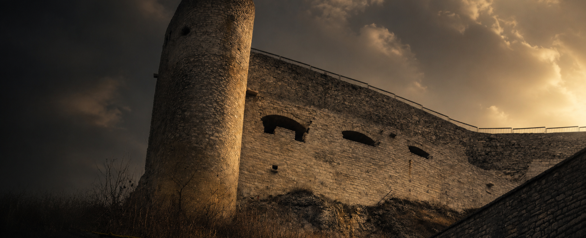 Eine mächtige Steinfestung mit rundem Turm erhebt sich unter einem dramatisch bewölkten Himmel. Das warme Abendlicht beleuchtet die historischen Mauern der Burg Hohenneuffen.“
