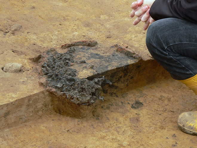 Ausgegrabenes Unterteil eines Rennofens in einer archäologischen Grabungsfläche; im Boden sind dunkle Verfärbungen und Reste der Ofenstruktur erkennbar.