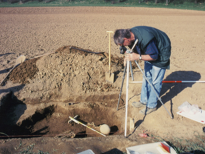 Ein Archäologe auf einem Feld der einen Fund vermisst man sieht im Grabungsloch einen runden Stein.