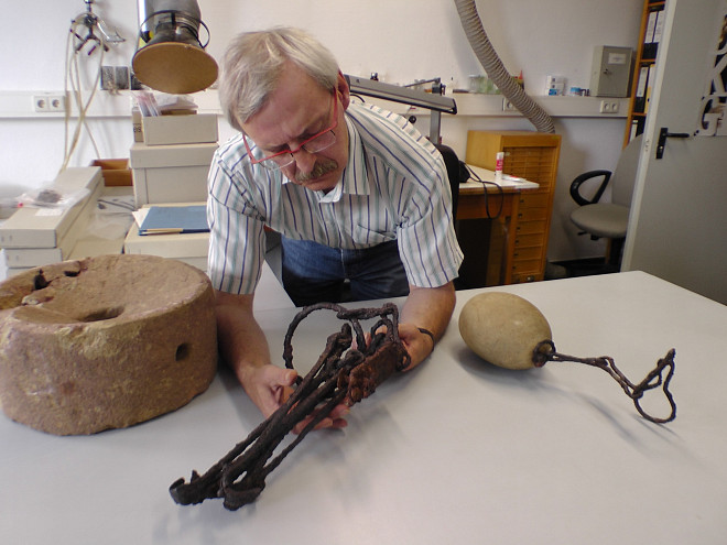 Ein Mühlstein, ein Eisengestänge und ein runder Stein auf dem Tisch eines Wissenschaftler, der das Eisengestänge in der Hand hält und nachdenklich betrachtet.