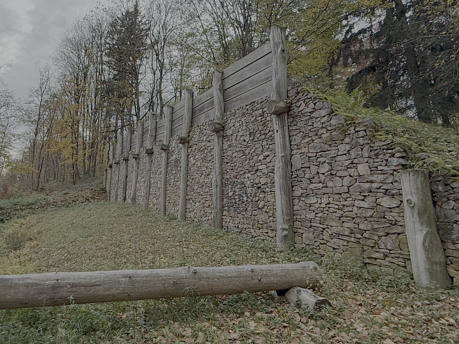 Rekonstruierte Posten Schlitzmauer am Donnersberg auf einer Wiese man sieht gut die Holzpfosten und die Trockenbauweise des Mauerwerks, obendrauf befinden sich befindet sich noch ein Palisadenwall aus Brettern
