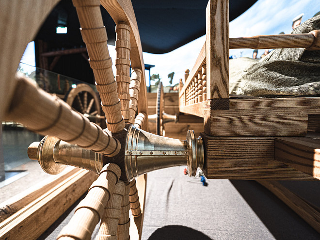 Nahaufnahme von unten eines keltischen hallstattzeitlichen Holzrades mit Drechselmustern. Zentriert sieht man die goldene metallbeschlagene Radnabe und Details des Wagens.