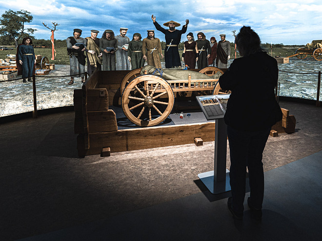 Ein Mensch als Schatten dargestellt, steht vor einer Texttafel und liest aufmerksam, die Informationen zu einem hölzern keltischen Wohnwagen im Hintergrund. Ein Panorama einer Grablege.