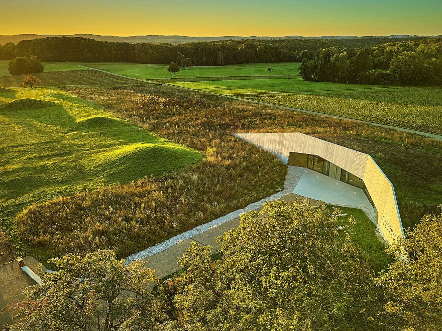 Ein moderner Betonbau bei Sonnenaufgang, umgeben von grüner Wiese, rechts im Bild keltische Grabhügel.