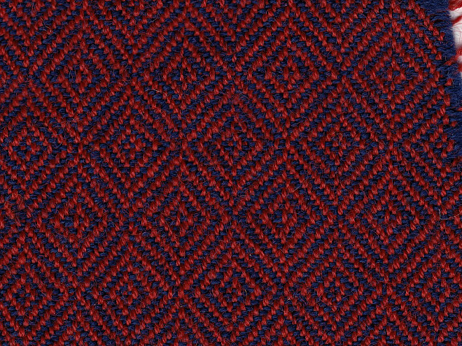 Nahaufnahme eines wollenen Diamantköpers mit rautenförmigem Webmuster, roten Kettfäden, blauen Schussfäden und einer sichtbaren Webkante.