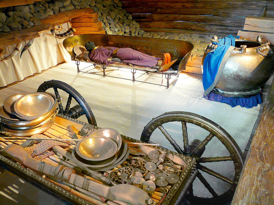 Rekonstruktion der Grabkammer des Hochdorfer Keltenfürsten im Keltenmuseum Hochdorf: Im Zentrum liegt eine Figur des Verstorbenen auf einer reich verzierten bronzenen Liege. Rechts steht ein großer Bronzekessel mit blauem Tuch, im Vordergrund ein zweirädriger Wagen mit bronzenen Schalen, Trinkgeschirr und weiteren Beigaben. Die hölzerne Grabkammer mit Steinmauerwerk ist warm beleuchtet und vermitt